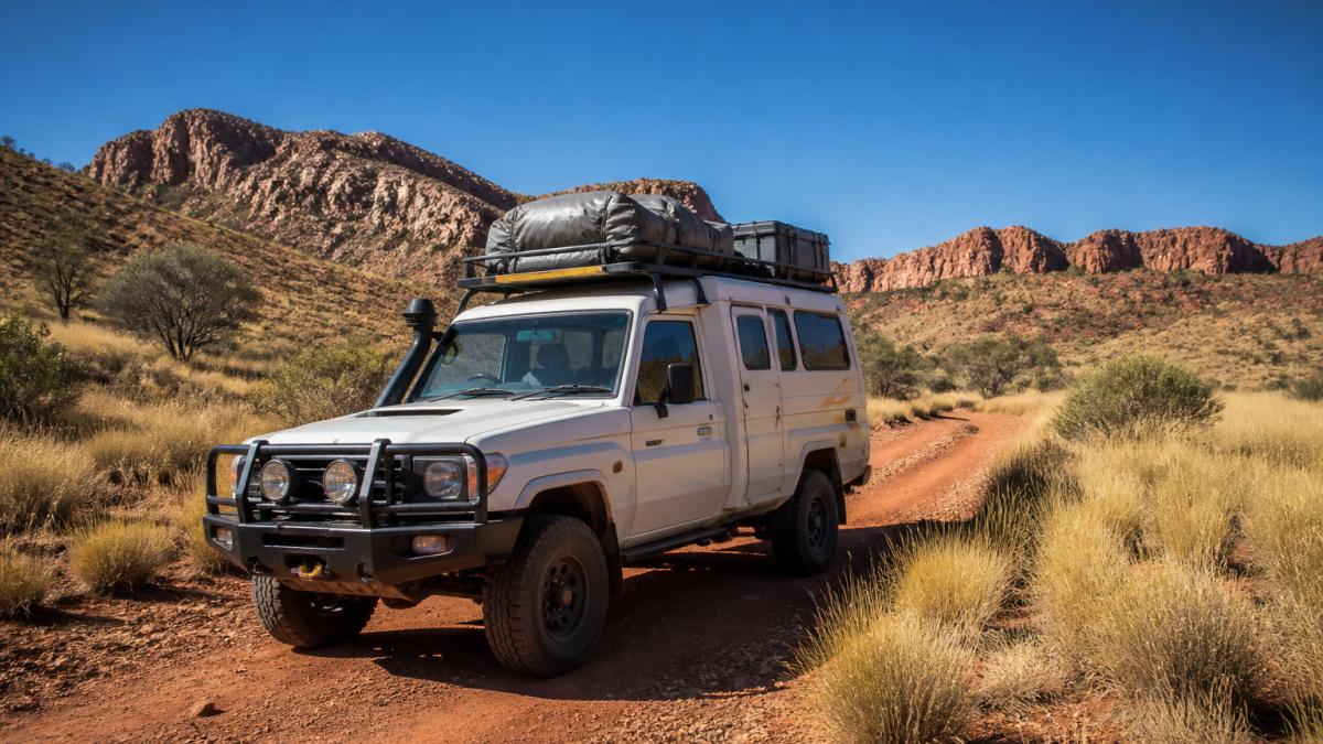 Well-equipped 4WD campervan parked on a red dirt track in remote outback terrain with spinifex grass, rocky ridges, and clear blue sky, showing the vehicle's rugged capability in isolated desert landscape