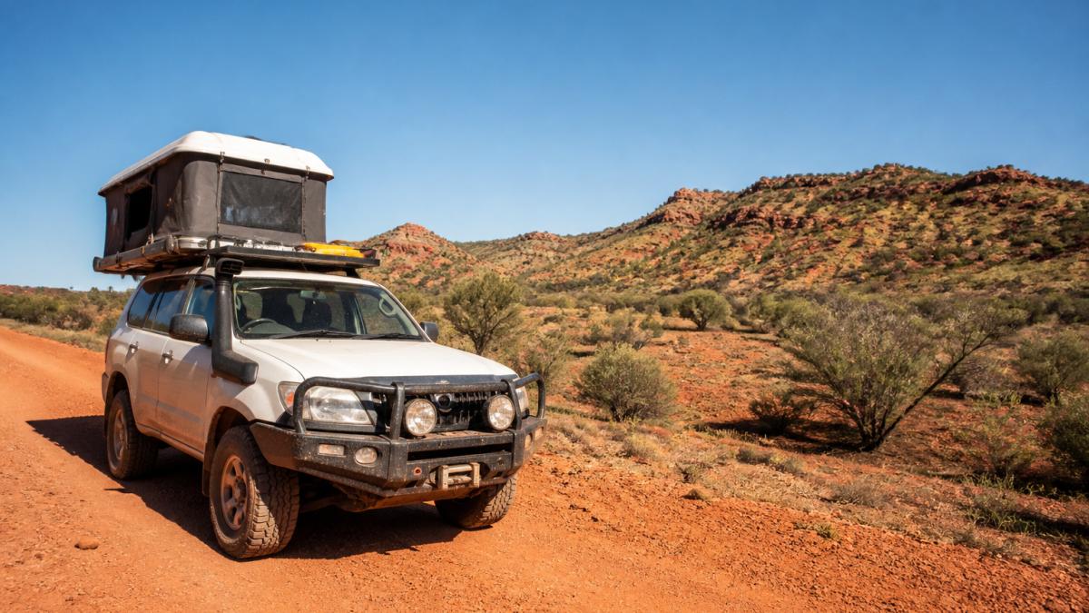 A 4WD camper parked on a red dirt unsealed road with red rocky outback landscape, sparse vegetation, and clear sky in the background, showcasing the vehicle's off-road capability