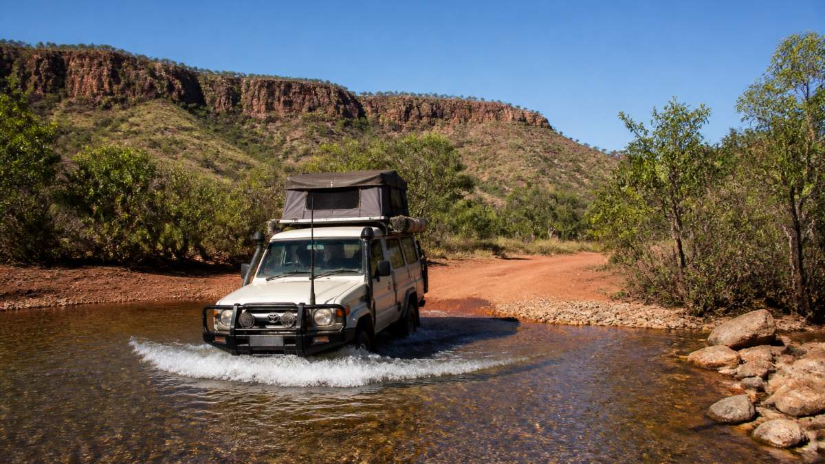 A 4WD vehicle with a roof-top tent crossing a shallow river on a remote red dirt track in the Kimberley, rugged escarpment country and a clear blue sky behind it
