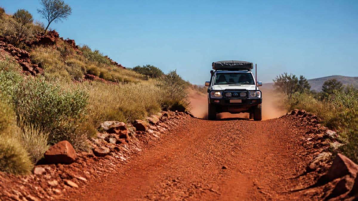 A high-clearance 4WD camper driving along a red dirt road in Karijini National Park, with spinifex and rocky outcrops on either side and dust trailing behind the vehicle under a clear outback sky
