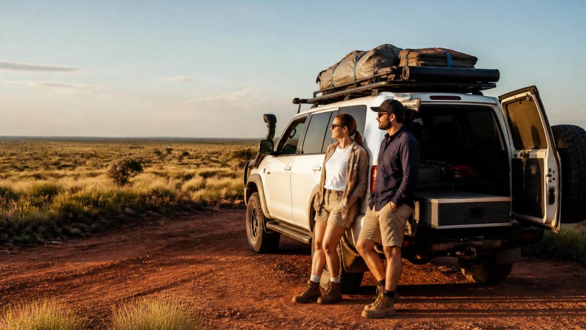A couple standing beside a rugged 4WD camper on a red dirt track with spinifex and low scrub stretching to the horizon, roof rack loaded with gear, late afternoon light